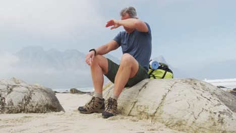 Hombre-Cansado-Excursionista-Mayor-Con-Mochila-Usando-Smartwatch-Mientras-Está-Sentado-En-Una-Caminata-De-Roca-En-La-Playa.
