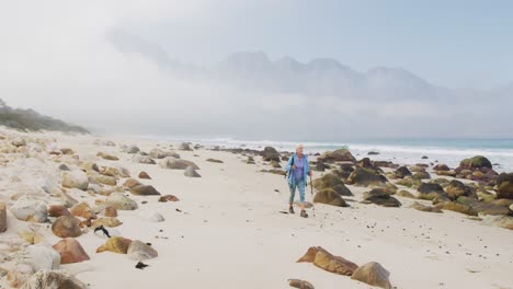 Senior-hiker-woman-with-backpack-and-hiking-poles-walking-while-hiking-on-the-beach.
