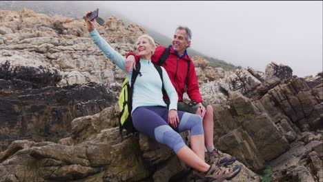 Senior-hiker-couple-with-backpacks-sitting-on-the-rocks-and-taking-a-selfie-on-smartphone