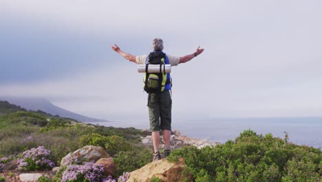 Rear-view-of-senior-hiker-man-standing-with-his-arms-wide-open-standing-on-a-rock