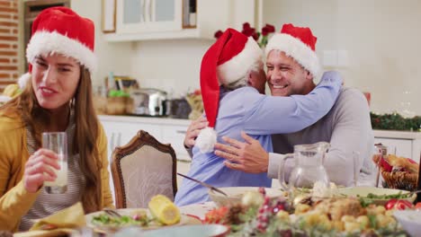 Kaukasische-ältere-Frau-Mit-Weihnachtsmütze-Umarmt-Ihren-Sohn,-Während-Sie-Am-Esstisch-Sitzt-Und-Das-Mittagessen-Genießt