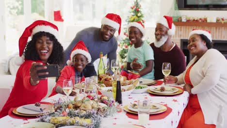 Familia-Afroamericana-Con-Gorros-De-Papá-Noel-Tomando-Un-Selfie-En-Un-Teléfono-Inteligente-Mientras-Está-Sentado-En-La-Mesa-Del-Comedor.