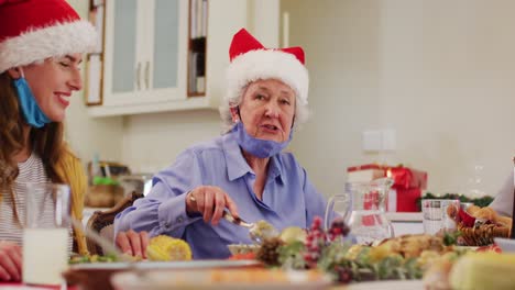 Caucasian-senior-woman-wearing-santa-hat-and-face-mask-around-her-neck-smiling-while-sitting-on-dini