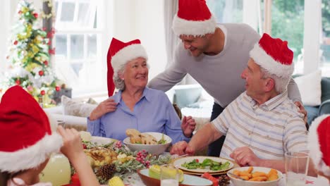 Kaukasisches-älteres-Paar-Mit-Weihnachtsmützen-Umarmt-Seinen-Sohn-Und-Genießt-Gemeinsam-Das-Mittagessen-Im-Sitzen