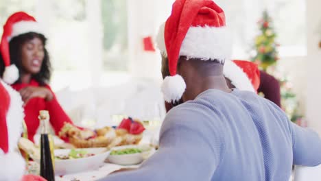 Portrait-of-african-american-man-in-santa-hat-smiling-while-sitting-on-dining-table-having-lunch-tog