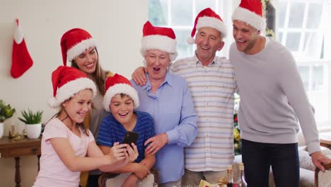 Kaukasische-Familie-Trägt-Weihnachtsmützen-Und-Nutzt-Gemeinsam-Ihr-Smartphone-Im-Wohnzimmer-Zu-Hause