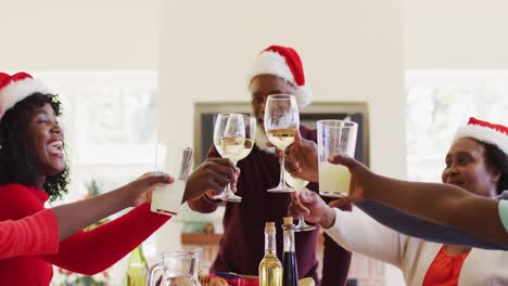 Afroamerikanische-Familie-In-Weihnachtsmützen-Toastet,-Während-Sie-Am-Esstisch-Sitzt-Und-Gemeinsam-Zu-Mittag-Isst-A