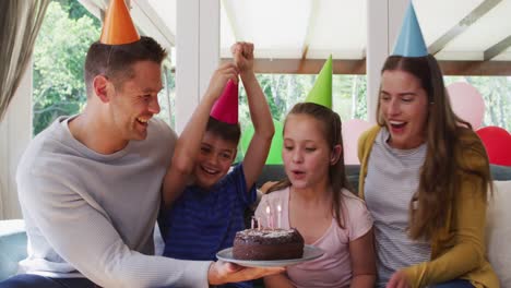 Portrait-of-caucasian-girl-in-party-hat-blowing-candles-on-birthday-cake-while-family-smiling-and-wa