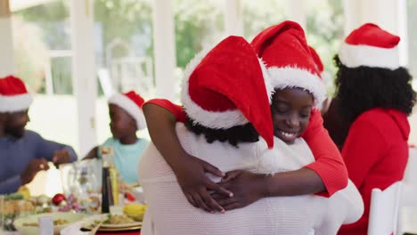 African-american-girl-in-santa-hat-hugging-his-grandmother-while-sitting-on-dining-table-having-lunc