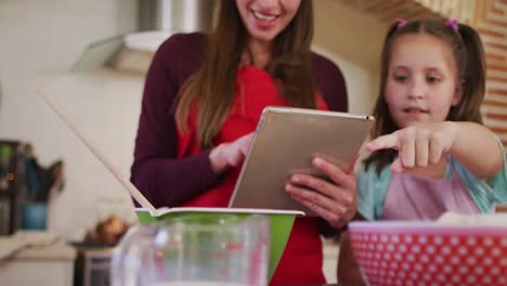 Caucasian-mother-and-daughter-wearing-aprons-using-digital-tablet-while-baking-in-the-kitchen-at-hom