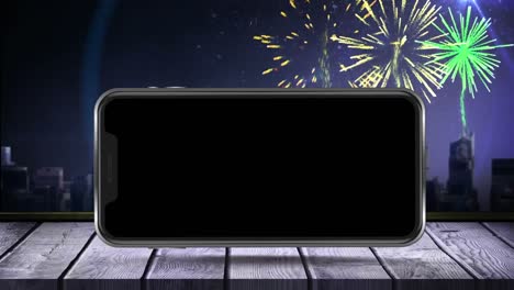 Digital-animation-of-smartphone-on-wooden-surface-against-fireworks-exploding-over-cityscape