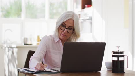 Senior-woman-wearing-glasses-taking-notes-and-using-laptop-while-working-from-home