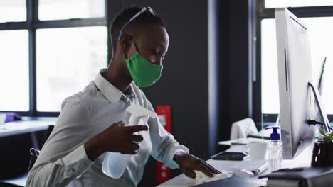 African-american-woman-wearing-face-mask-cleaning-her-desk-with-disinfectant-at-modern-office