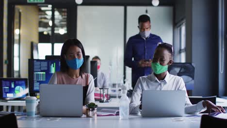 Diverse-female-colleagues-wearing-face-masks-greeting-each-other-by-touching-elbows-at-modern-office