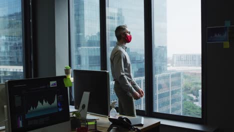 Thoughtful-caucasian-man-wearing-face-mask-looking-out-of-window-at-modern-office
