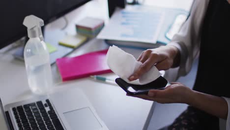 Mid-section-of-woman-cleaning-smartphone-with-tissue-at-modern-office
