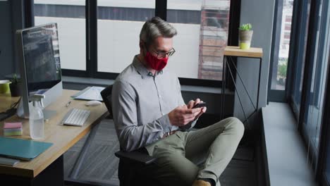 Kaukasischer-Mann-Mit-Gesichtsmaske-Spricht-Auf-Dem-Smartphone,-Während-Er-Auf-Seinem-Schreibtisch-Im-Modernen-Büro-Sitzt