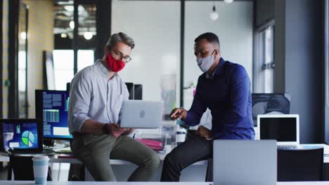 Diverse-male-colleagues-wearing-face-masks-discussing-over-laptop-at-modern-office