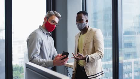 Diverse-male-office-colleagues-wearing-face-masks-discussing-over-smartphone-at-modern-office