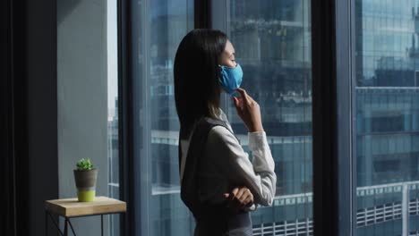 Thoughtful-asian-woman-wearing-face-mask-looking-out-of-window-at-modern-office