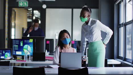 African-american-woman-and-asian-woman-wearing-face-masks-discussing-over-laptop-at-modern-office