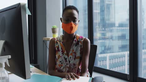 African-american-woman-wearing-face-mask-reading-file-of-documents-while-sitting-on-her-desk-at-mode
