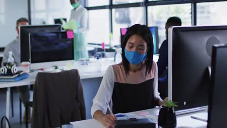 Asiatische-Frau-Mit-Gesichtsmaske-Macht-Sich-Notizen,-Während-Sie-Auf-Ihrem-Schreibtisch-Im-Modernen-Büro-Sitzt
