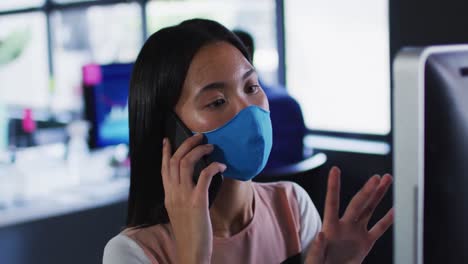 Asiatische-Frau-Mit-Gesichtsmaske-Spricht-Auf-Dem-Smartphone,-Während-Sie-Auf-Ihrem-Schreibtisch-Im-Modernen-Büro-Sitzt