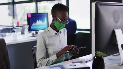 Afroamerikanische-Frau-Mit-Gesichtsmaske-Benutzt-Computer-Und-Digitales-Tablet,-Während-Sie-Auf-Ihrem-Schreibtisch-Sitzt