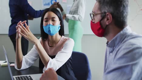 Diverse-male-and-female-office-colleagues-wearing-face-masks-discussing-over-laptop-at-modern-office