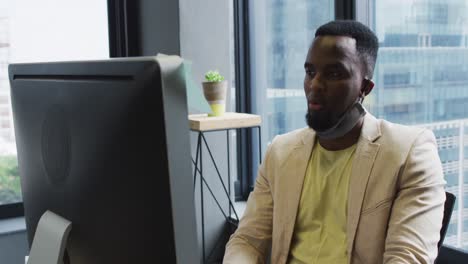 African-american-man-with-face-mask-around-his-neck-using-computer-while-sitting-on-his-desk-at-mode