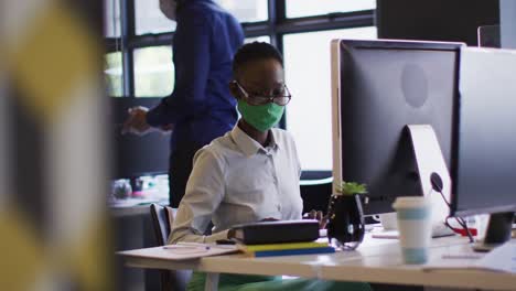 Afroamerikanische-Frau-Mit-Gesichtsmaske-Und-Computer,-Während-Sie-Auf-Ihrem-Schreibtisch-Im-Modernen-Büro-Sitzt