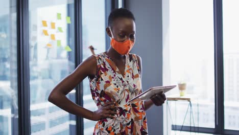 Portrait-of-african-american-woman-wearing-face-mask-using-digital-tablet-at-modern-office