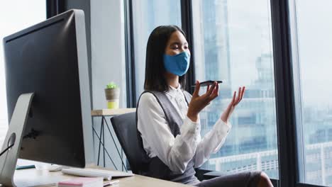 Asiatische-Frau-Mit-Gesichtsmaske-Spricht-Auf-Dem-Smartphone,-Während-Sie-Auf-Ihrem-Schreibtisch-Im-Modernen-Büro-Sitzt