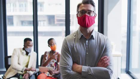 Portrait-of-caucasian-man-wearing-face-mask-standing-with-his-arms-crossed-at-modern-office