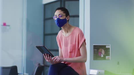 Portrait-of-caucasian-woman-wearing-face-mask-using-digital-tablet-at-modern-office