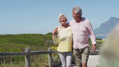 Happy-senior-caucasian-couple-with-arms-around-each-other-walking-on-path-leading-to-the-beach