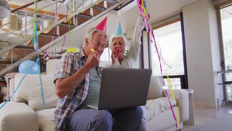 Älteres-Kaukasisches-Paar-Bläst-Partygebläse-Und-Feiert-Geburtstag,-Während-Es-Einen-Videoanruf-Auf-Dem-Laptop-Führt