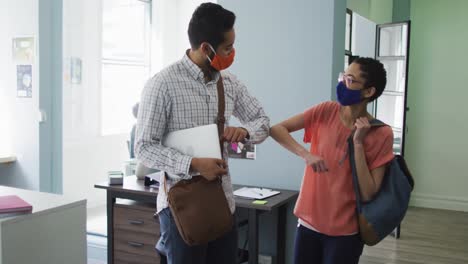 Diverse-male-and-female-colleague-wearing-face-masks-greeting-each-other-by-touching-elbows-at-moder