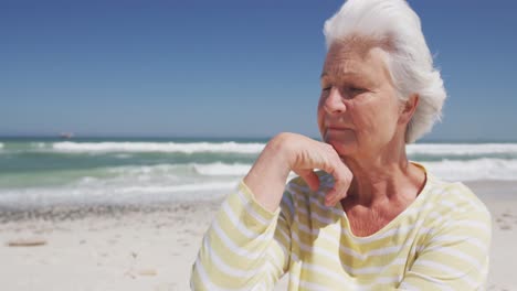 Thoughtful-senior-caucasian-woman-with-hand-on-chin-sitting-on-the-beach