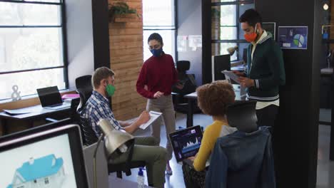 Diverse-colleagues-wearing-face-masks-with-electronic-devices-discussing-together-at-modern-office