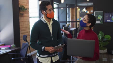 Diverse-male-and-female-colleagues-wearing-face-masks-using-laptop-digital-tablet-discussing-at-mode