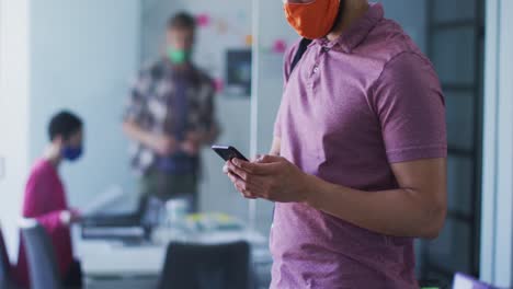 Mixed-race-man-wearing-face-mask-using-smartphone-in-office