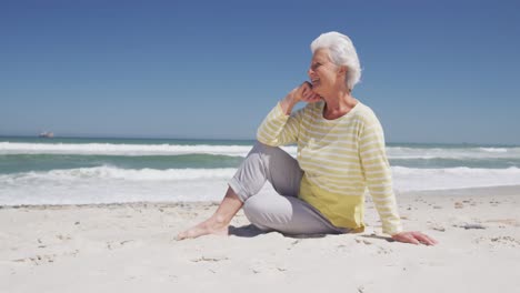 Happy-senior-caucasian-woman-with-hand-on-chin-sitting-on-the-beach-enjoying-the-view