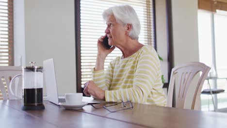 Ältere-Kaukasische-Frau-Mit-Laptop,-Die-Zu-Hause-Auf-Dem-Smartphone-Spricht