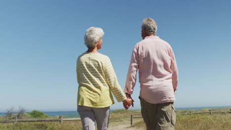 Senior-caucasian-couple-holding-hands-and-walking-on-path-leading-to-the-beach