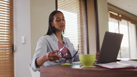 Mixed-race-woman-using-laptop-writing-in-notebook-drinking-coffee-working-from-home