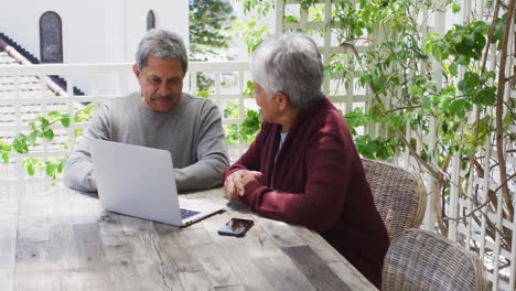 Happy-senior-mixed-race-couple-sitting-in-garden-using-laptop