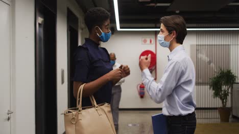 Diverse-business-people-wearing-face-masks-greeting-in-corridor-using-elbows