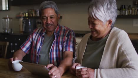 Happy-senior-mixed-race-couple-having-coffee-laughing-in-kitchen